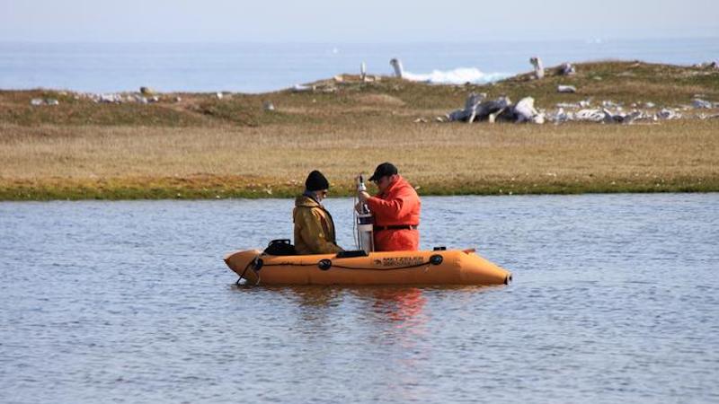 Sampling crew collecting a sediment core at PaJs-13, a Thule-Inuit site on Somerset Island, Nunavut, with the remnants of whalebone houses visible in the background. Photo credit: Jules Blais, University of Ottawa